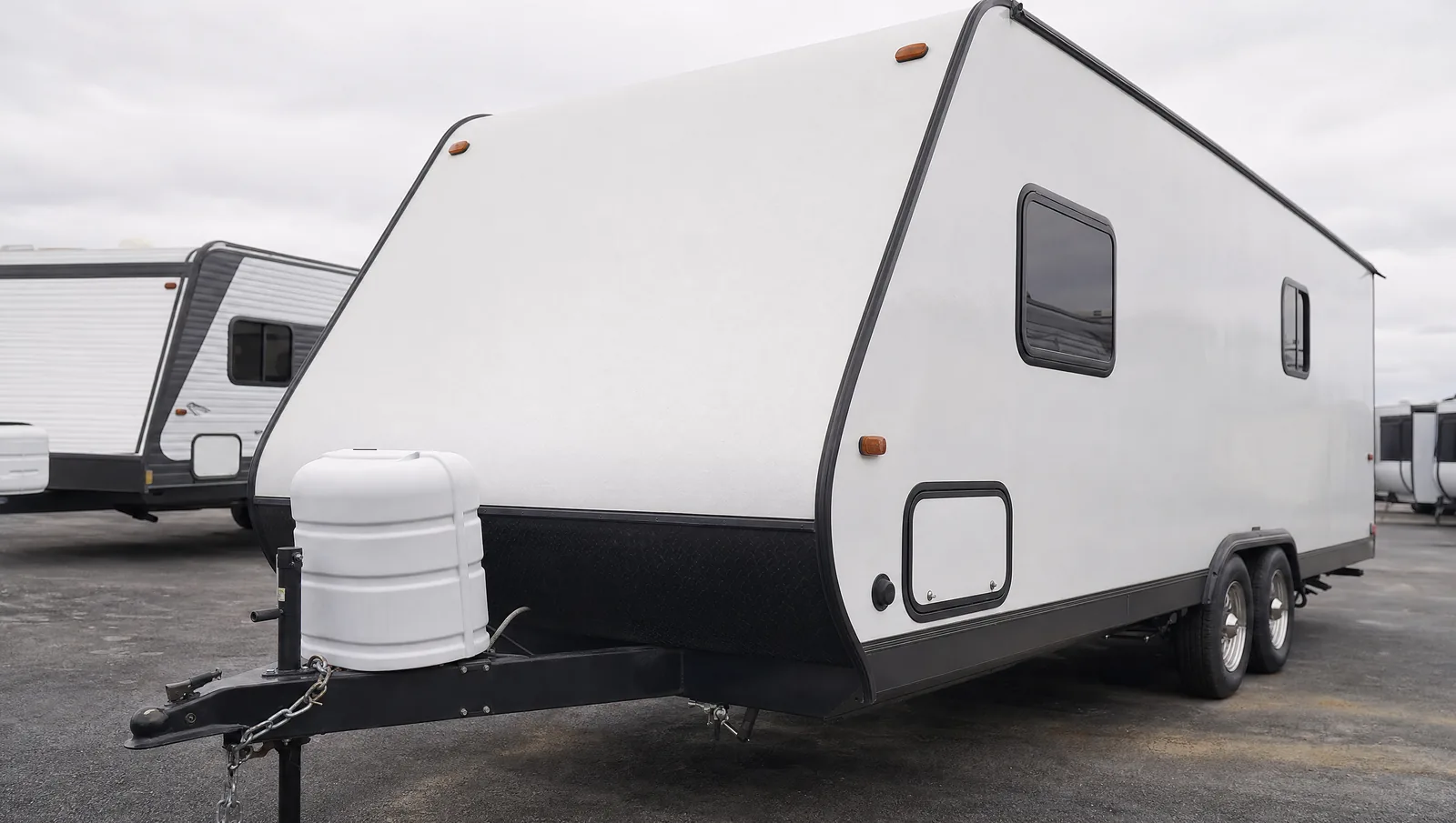 A white travel trailer on a dealership lot, photographed from the front three-quarter angle with its A-frame tongue, coupler and propane tank cover in the foreground.