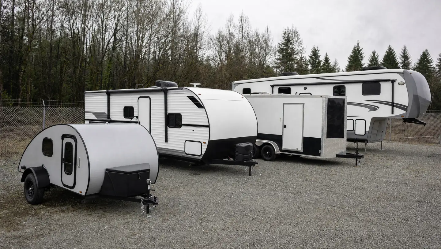 Four trailers parked in a storage yard in order of size: a small teardrop, a mid-size travel trailer, a tandem-axle enclosed cargo trailer and a large fifth-wheel RV.